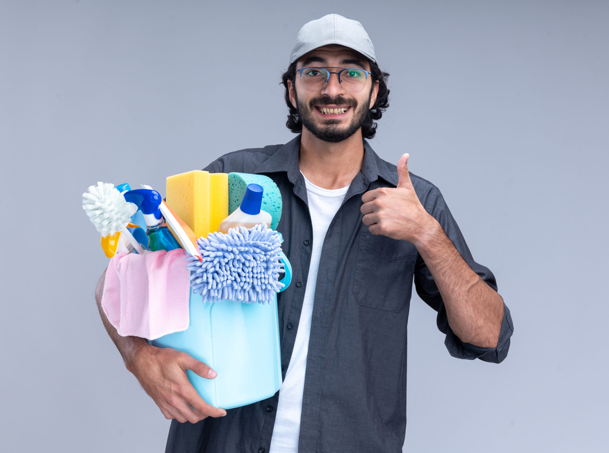 smiling young handsome cleaning guy wearing t-shirt and cap holding bucket of cleaning tools showing thumb up isolated on white background